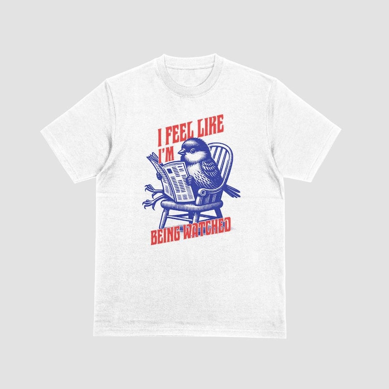 May include: White t-shirt with a blue and red graphic of a bird reading a newspaper in a chair. The text "I FEEL LIKE I'M BEING WATCHED" is printed above and below the bird illustration.