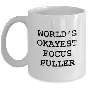 May include: White ceramic coffee mug with black text that reads "World's Okayest Focus Puller"