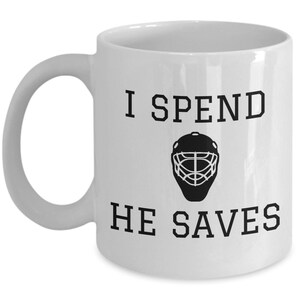 May include: White ceramic coffee mug with black text that reads "I spend, he saves" and a black silhouette of a hockey goalie mask.