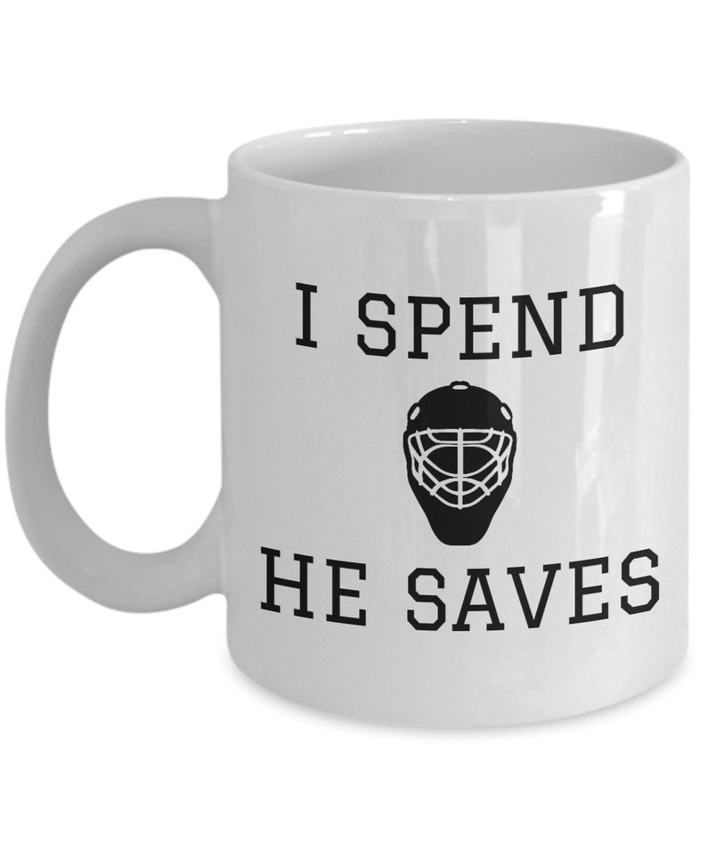 May include: White ceramic coffee mug with black text that reads "I spend, he saves" and a black silhouette of a hockey goalie mask.