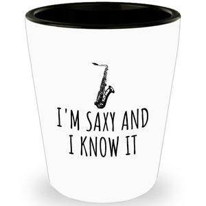 Puede incluir: Vaso de chupito blanco con borde negro y el texto "I'M SAXY AND I KNOW IT" en negro. Un gráfico de saxofón en blanco y negro está encima del texto.