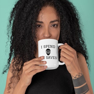 May include: White ceramic mug with black text that reads "I spend all my saves". A hockey goalie mask graphic is between the words "spend" and "saves". A person is holding the mug and drinking from it.