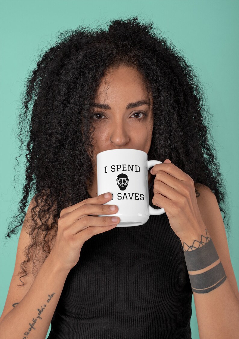 May include: White ceramic mug with black text that reads "I spend all my saves". A hockey goalie mask graphic is between the words "spend" and "saves". A person is holding the mug and drinking from it.