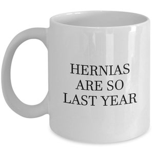 May include: White ceramic mug with a curved handle. The mug features the text "HERNIAS ARE SO LAST YEAR" in black, centered on the front. A humorous gift idea.