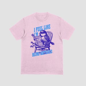 May include: Pale pink t-shirt featuring a blue illustration of a bird reading a newspaper, seated on a chair. The text "I FEEL LIKE I'M BEING WATCHED" is printed above and below the bird.