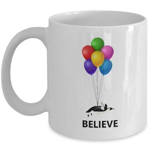 May include: White ceramic mug with a penguin holding onto colorful balloons. The text "BELIEVE" is printed on the mug.