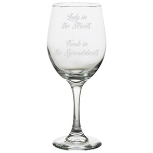 May include: A clear wine glass with a long stem and a wide bowl. The glass has the text "Lady in the Streets" and "Freak in the Spreadsheets" etched on the front. The glass is designed for drinking wine.