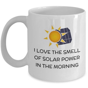 Pode incluir: Caneca de cerâmica branca com uma alça curva. A caneca apresenta um gráfico de um sol amarelo e um painel solar, com o texto "I LOVE THE SMELL OF SOLAR POWER IN THE MORNING."
