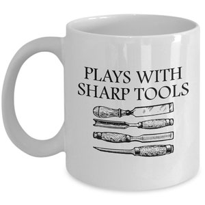 May include: White ceramic coffee mug with black text that reads "Plays with sharp tools". The mug features a black and white illustration of four different woodworking tools.