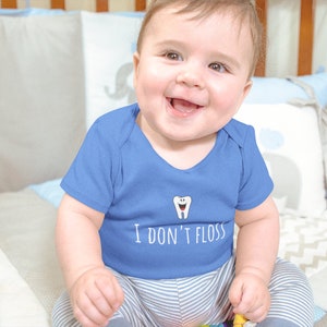 Peut inclure: Un b&eacute;b&eacute; portant un body bleu avec un dessin de dent blanche et le texte "Je ne passe pas de fil dentaire". Le b&eacute;b&eacute; est assis sur une couverture &agrave; motifs blancs et gris.
