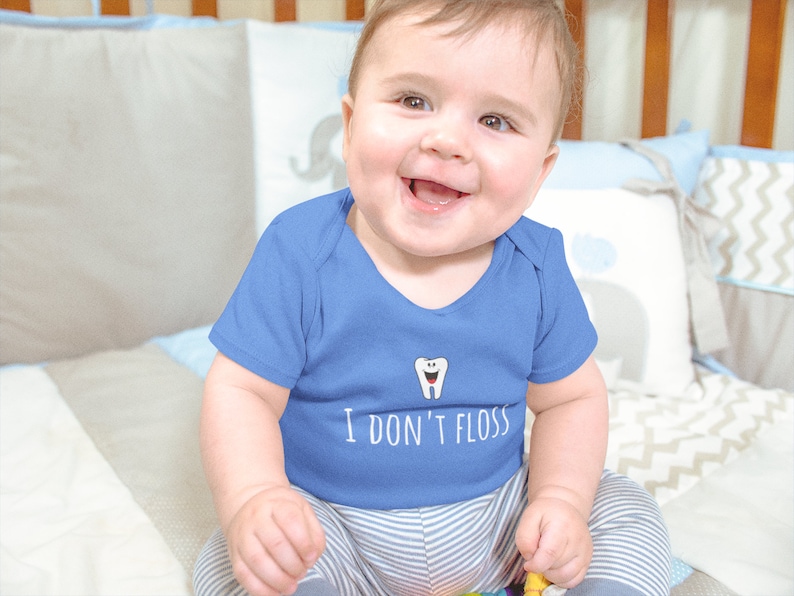 Peut inclure: Un b&eacute;b&eacute; portant un body bleu avec un dessin de dent blanche et le texte "Je ne passe pas de fil dentaire". Le b&eacute;b&eacute; est assis sur une couverture &agrave; motifs blancs et gris.