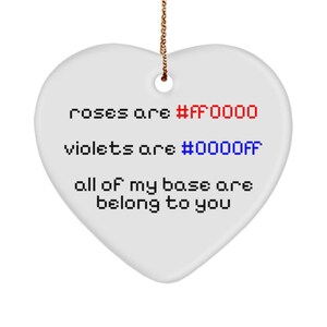 Puede incluir: Adorno blanco en forma de corazón con una cuerda dorada. El adorno tiene el texto "roses are #FF0000, violets are #0000FF, all of my base are belong to you" en una fuente pixelada. El texto es en negro, rojo y azul.