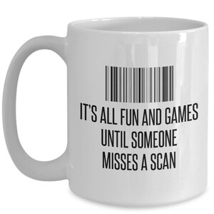 May include: White ceramic mug with a black barcode graphic and the text "IT'S ALL FUN AND GAMES UNTIL SOMEONE MISSES A SCAN". The mug has a curved handle and a cylindrical shape.