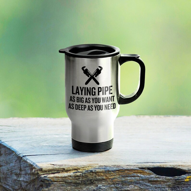May include: A silver travel mug with a black lid and handle. The mug has a black graphic of two crossed wrenches and the text "Laying Pipe As Big As You Want As Deep As You Need".