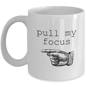 May include: White ceramic coffee mug with black text that reads "pull my focus" and a black and white illustration of a hand pointing to the right.