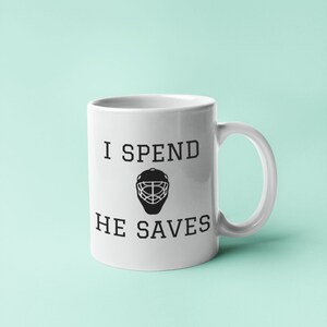 May include: White ceramic coffee mug with black text that reads "I spend, he saves" and a black hockey goalie mask graphic.