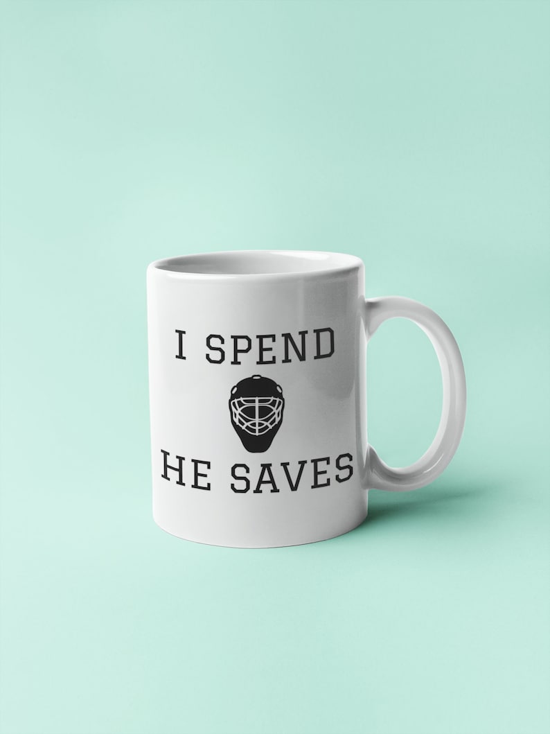 May include: White ceramic coffee mug with black text that reads "I spend, he saves" and a black hockey goalie mask graphic.