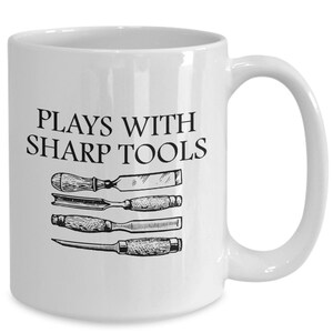 May include: White ceramic coffee mug with a black inscription that reads "Plays with Sharp Tools".  Four black and white illustrations of chisels are displayed below the text.