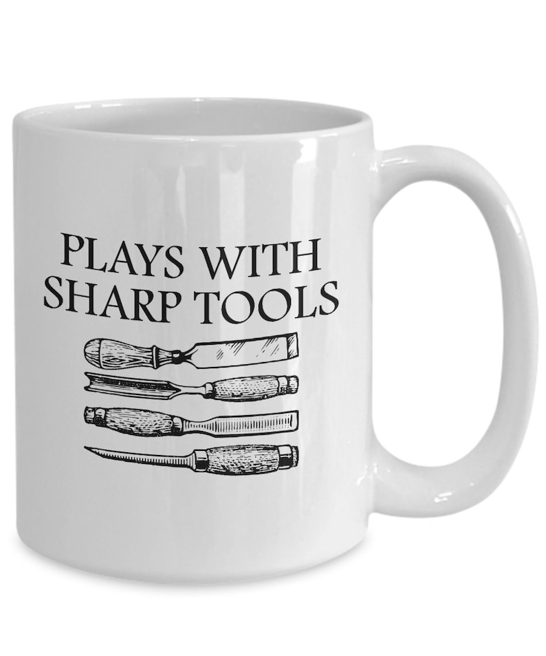 May include: White ceramic coffee mug with a black inscription that reads "Plays with Sharp Tools".  Four black and white illustrations of chisels are displayed below the text.