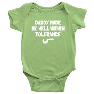 May include: A light green baby onesie with the text "DADDY MADE ME WELL WITHIN TOLERANCE" in white. A white wrench graphic is below the text. The onesie has short sleeves and snaps at the bottom.