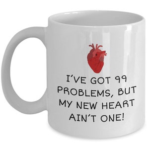 Pode incluir: Caneca de café de cerâmica branca com uma ilustração de um coração anatômico vermelho e o texto "Tenho 99 problemas, mas meu novo coração não é um!"