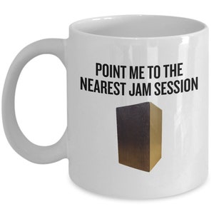 May include: White ceramic mug with the text "POINT ME TO THE NEAREST JAM SESSION" in black. A brown and gold cajon drum graphic is below the text. The mug has a curved handle.
