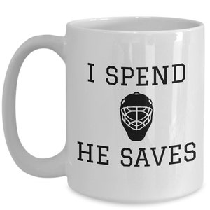 May include: White ceramic coffee mug with a black graphic that says "I spend, he saves" with a hockey goalie mask in between the words.
