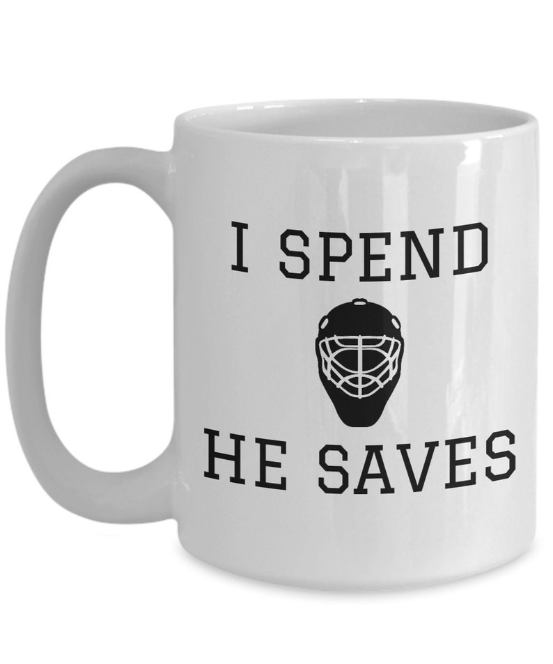 May include: White ceramic coffee mug with a black graphic that says "I spend, he saves" with a hockey goalie mask in between the words.