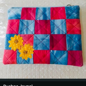 K&ouml;nnte beinhalten: Eine gesteppte Stofftasche mit Rei&szlig;verschluss. Die Tasche ist aus blauem und rotem Stoff mit Patchwork-Muster. Zwei gestickte gelbe Blumen befinden sich auf der Vorderseite der Tasche.