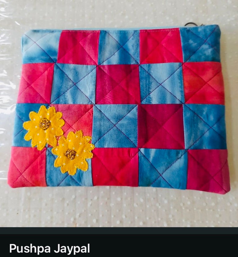 K&ouml;nnte beinhalten: Eine gesteppte Stofftasche mit Rei&szlig;verschluss. Die Tasche ist aus blauem und rotem Stoff mit Patchwork-Muster. Zwei gestickte gelbe Blumen befinden sich auf der Vorderseite der Tasche.