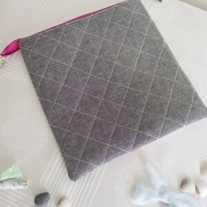 K&ouml;nnte beinhalten: Eine graue gesteppte Rei&szlig;verschlusstasche mit einem rosa Rei&szlig;verschluss. Die Tasche liegt auf einer wei&szlig;en Oberfl&auml;che mit Muscheln und einem kleinen blauen Stoffst&uuml;ck.