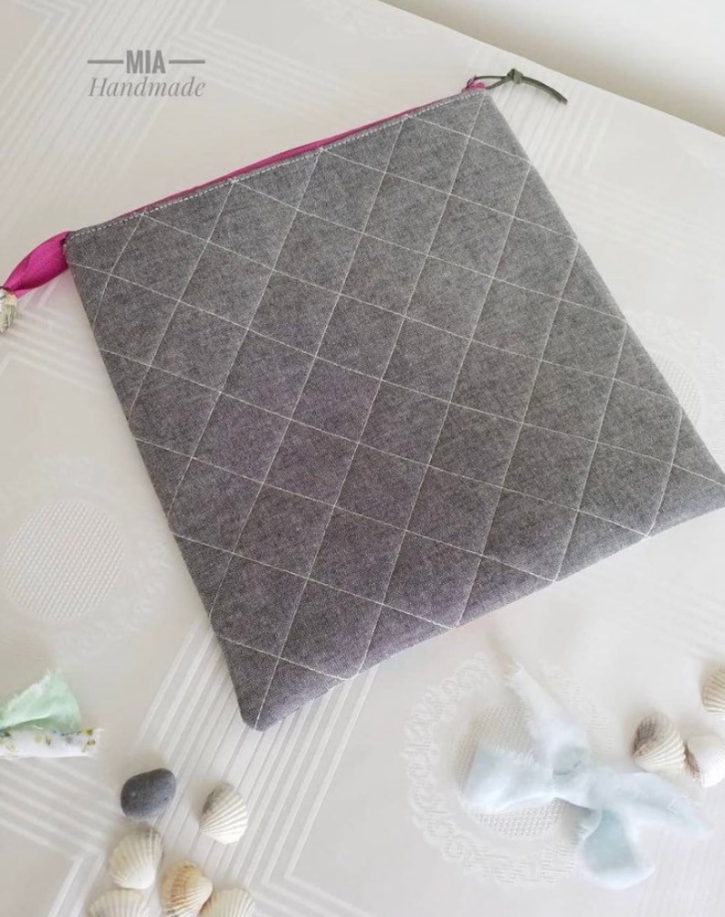 K&ouml;nnte beinhalten: Eine graue gesteppte Rei&szlig;verschlusstasche mit einem rosa Rei&szlig;verschluss. Die Tasche liegt auf einer wei&szlig;en Oberfl&auml;che mit Muscheln und einem kleinen blauen Stoffst&uuml;ck.