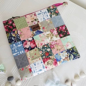 May include: A quilted pouch with a patchwork design featuring floral and geometric patterns in shades of pink, blue, green, and gray. The pouch has a zipper closure and a pink pull tab. The pouch is labeled "CHARMING QUILTED POUCH" and "MIA Handmade".