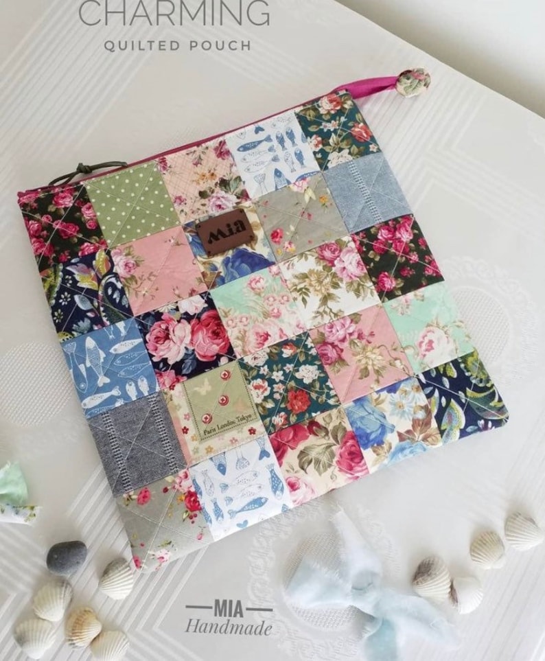 K&ouml;nnte beinhalten: Eine gesteppte Tasche mit Patchwork-Design, das florale und geometrische Muster in Rosa, Blau, Gr&uuml;n und Grau zeigt. Die Tasche hat einen Rei&szlig;verschluss und einen rosa Zugverschluss. Die Tasche ist mit "CHARMING QUILTED POUCH" und "MIA Handmade" beschriftet.