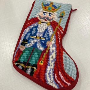 Puede incluir: Calcetín navideño de punto de aguja con diseño de rey cascanueces. El calcetín tiene un borde y cremallera rojos, con un fondo azul claro. El rey lleva una corona dorada, capa roja y chaqueta azul. Una decoración navideña única.