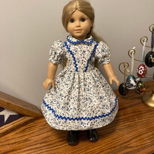 Könnte beinhalten: Eine Puppe, die ein weißes Kleid mit blauem Blumendruck und blauem Besatz trägt. Das Kleid hat einen blauen Kragen und Puffärmel. Die Puppe hat blonde Haare und blaue Augen.