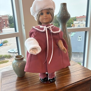 May include: A doll with light brown hair and blue eyes wearing a dusty rose cape with a white faux fur collar and cuffs, a matching white faux fur beret, and a white ribbon tie. The doll is dressed in matching dusty rose pants and black shoes.