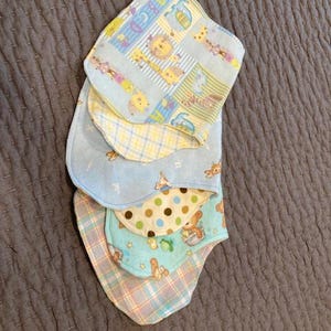 May include: A stack of four baby bibs with different patterns. The top bib is blue with a yellow and white checked pattern and features a variety of animal illustrations. The second bib is blue with a yellow and white checked pattern. The third bib is brown with white polka dots. The fourth bib is blue with a yellow and white checked pattern and features a variety of animal illustrations.