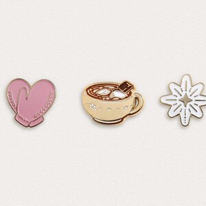 May include: Three gold enamel pins with a winter theme. The first pin is a pair of pink mittens. The second pin is a cup of hot chocolate with marshmallows and a chocolate square on top. The third pin is a white starburst.
