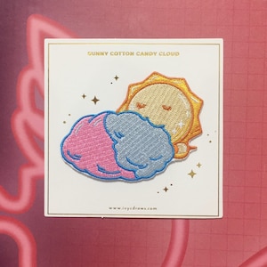 Puede incluir: Un sol amarillo con los ojos cerrados está durmiendo sobre una nube de algodón de azúcar rosa y azul. El parche tiene el texto "SUNNY COTTON CANDY CLOUD" y "www.ivycdraws.com".