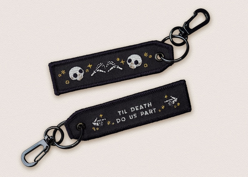 May include: Two black keychains with gold embroidery. One keychain features two skeletons holding hands with the text "Til Death Do Us Part". The other keychain features two skeletons holding hands with a skull in the middle.