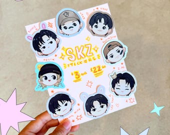 May include: A hand holding a white card with colorful stickers of cartoon characters. The stickers have the text "SKZ STICKERS" and feature various characters with different hairstyles and accessories. The background has star and heart shapes.