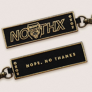 May include: Two black fabric keychains with gold embroidery. The first keychain has the text "NOOTHX" and a small cartoon figure holding a sign that says "NOPE". The second keychain has the text "NOPE. NO THANKS".