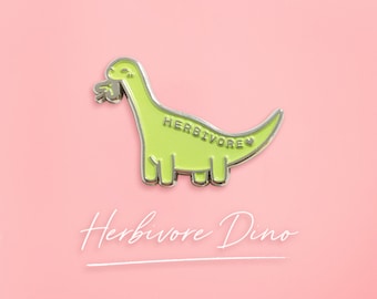 May include: A green dinosaur pin with a silver outline. The dinosaur is eating a green leaf and has the word "HERBIVORE" written on its side. The text "Herbivore Dino" is written below the pin.