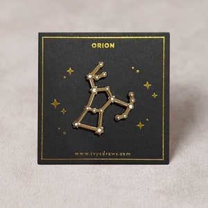 Puede incluir: Pin de color dorado con un contorno blanco de la constelación de Orión sobre un fondo negro. El pin está en una tarjeta negra con el texto "ORION" y "www.ivycdraws.com".