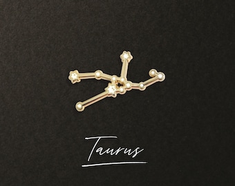 Puede incluir: Pin de la constelación de Tauro de color dorado con acentos de estrellas blancas sobre un fondo negro. La palabra "Taurus" está escrita en cursiva blanca debajo del pin.