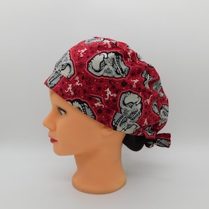 May include: A red scrub cap featuring a repeating pattern of grey elephant heads and white script A logos. The fabric has a dark red background with small black floral accents. The cap is tied at the back with a fabric knot and includes the text Pixie Scrub Cap.