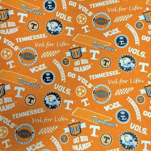 May include: An orange fabric featuring a repeating pattern of Tennessee Volunteers sports graphics, including footballs, helmets, the state outline, and text reading Tennessee, Vol for Life, Go Vols, and Go Big Orange in white and navy blue.