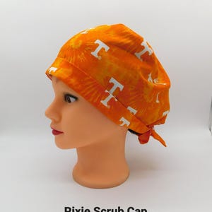 Könnte beinhalten: Eine orangefarbene und gelbe Pixie-OP-Haube mit weißen "T"-Logos. Die Haube ist hinten gebunden und wird auf einem Schaufensterkopf präsentiert. Der Text "Pixie Scrub Cap" ist unten im Bild aufgedruckt.