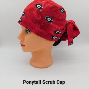 May include: A red tie-dye ponytail scrub cap featuring black and white "G" logos. The cap is tied in a bow at the back. The text "Ponytail Scrub Cap" is printed below the cap.